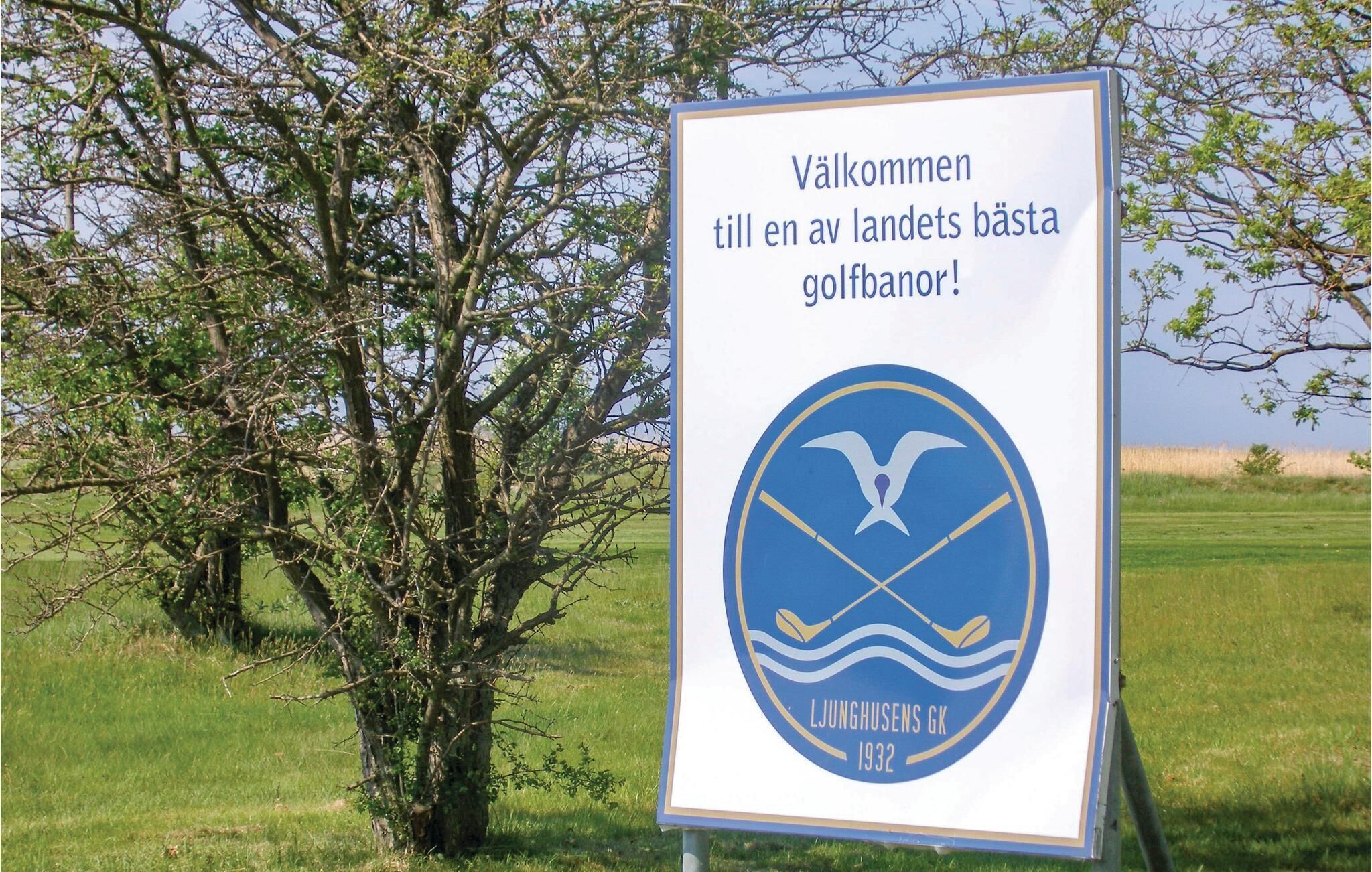 A sign with a logo and text welcoming visitors to a golf course, with a tree and grassy field in the background.