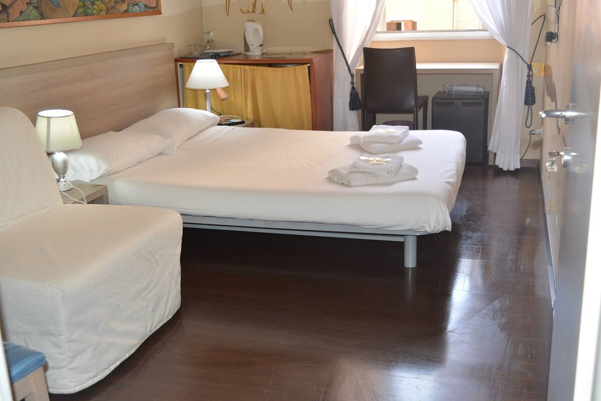 Une chambre d’hôtel avec un lit, une chaise, un bureau, une lampe et une fenêtre avec des rideaux.
