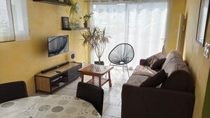 A living room with a sofa, TV, coffee table, and a chair. There are plants on shelves and a window with curtains. - Holiday house near sea and shops (La Baule)