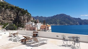 Terraza en la azotea con sillones y mesas, con vista a un pueblo costero con edificios históricos y un mar azul cristalino.