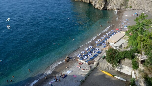 Un pueblo costero con una torre histórica, una playa con sombrillas y barcos en el agua.