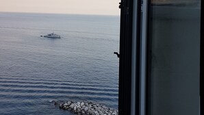 Vista desde una ventana sobre un mar tranquilo con un barco a lo lejos, una costa rocosa y un cielo despejado.