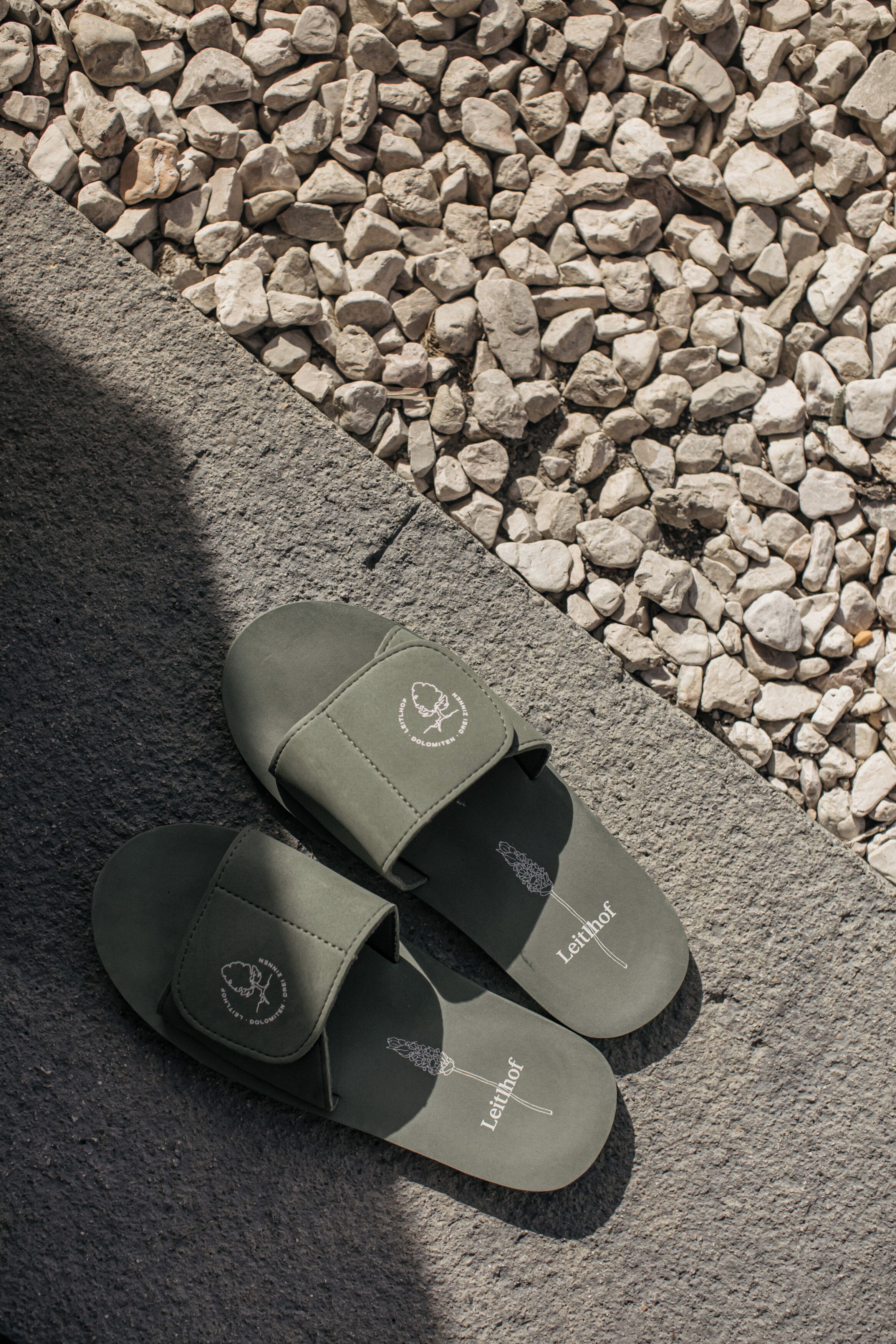 Sandalen mit dem Schriftzug "Leithof" auf der Sohle, auf einer Steinoberfläche neben einem Betonrand platziert.
