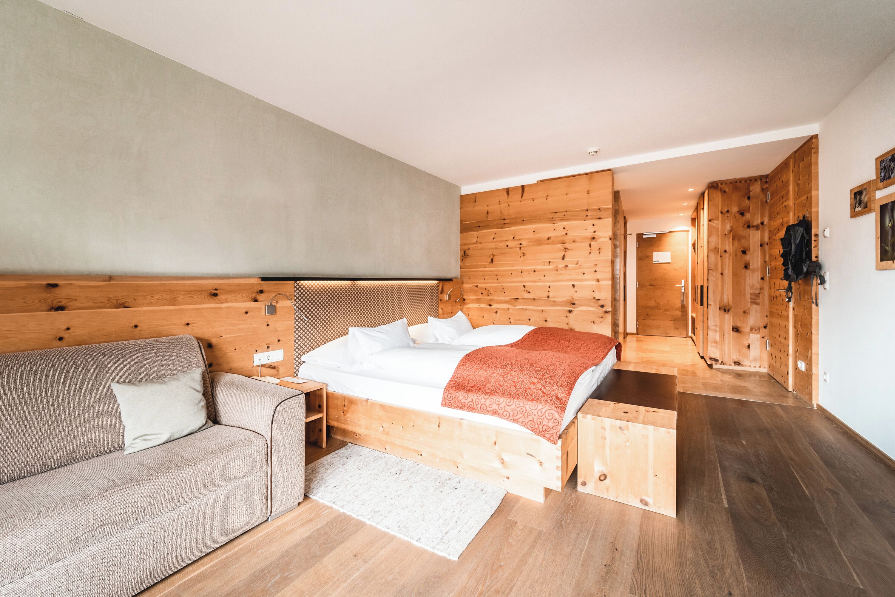 A modern bedroom with a wooden headboard, a bed with white linens and a red blanket, a grey sofa, and wooden flooring.