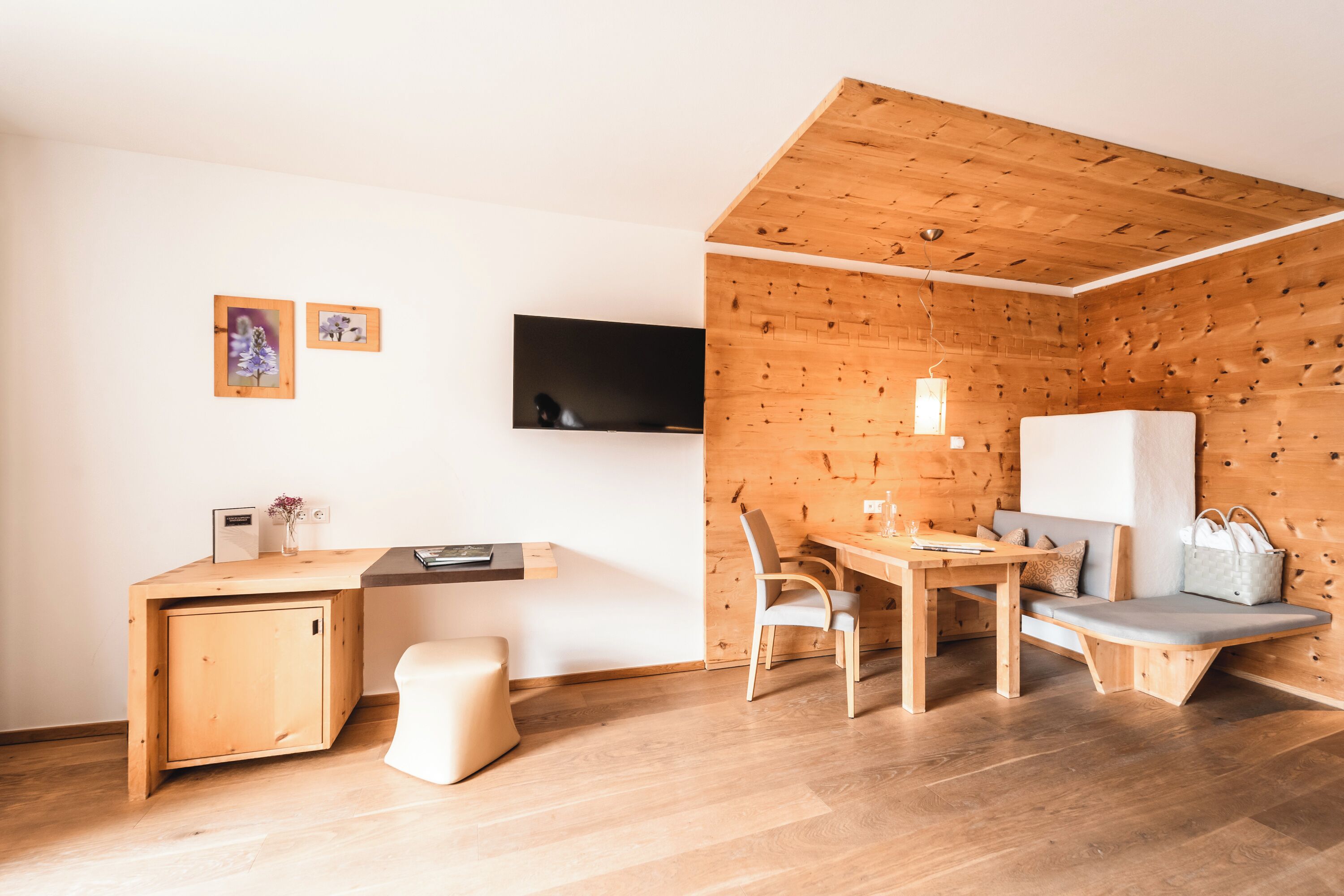 A modern, compact room with a wooden ceiling, a flat-screen TV mounted on the wall, a wooden desk with a chair, a bench with a cushion, and a small cabinet.