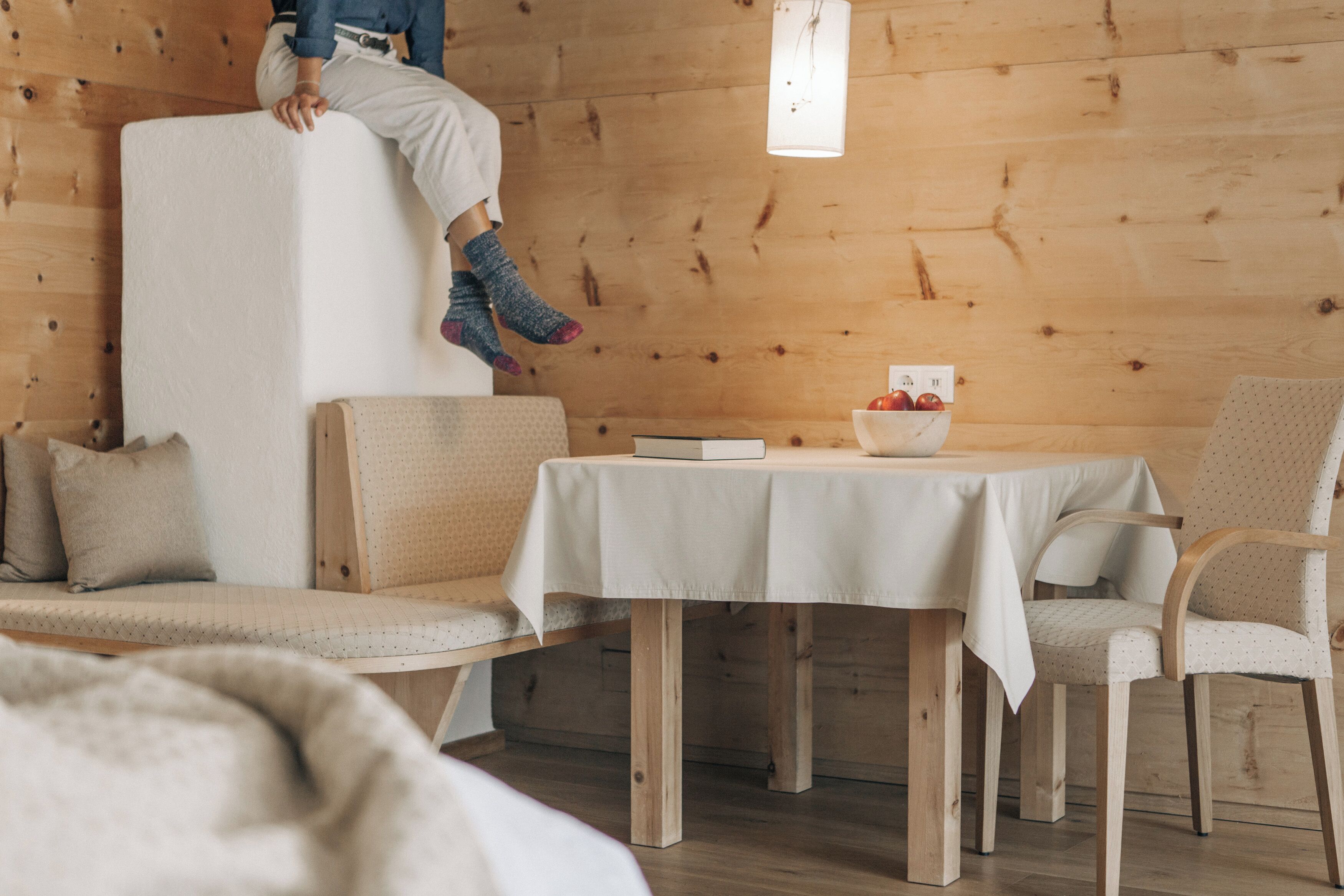 A cozy room with a wooden wall, a white sofa, a small table with a bowl of fruit, and a person sitting on a white object.