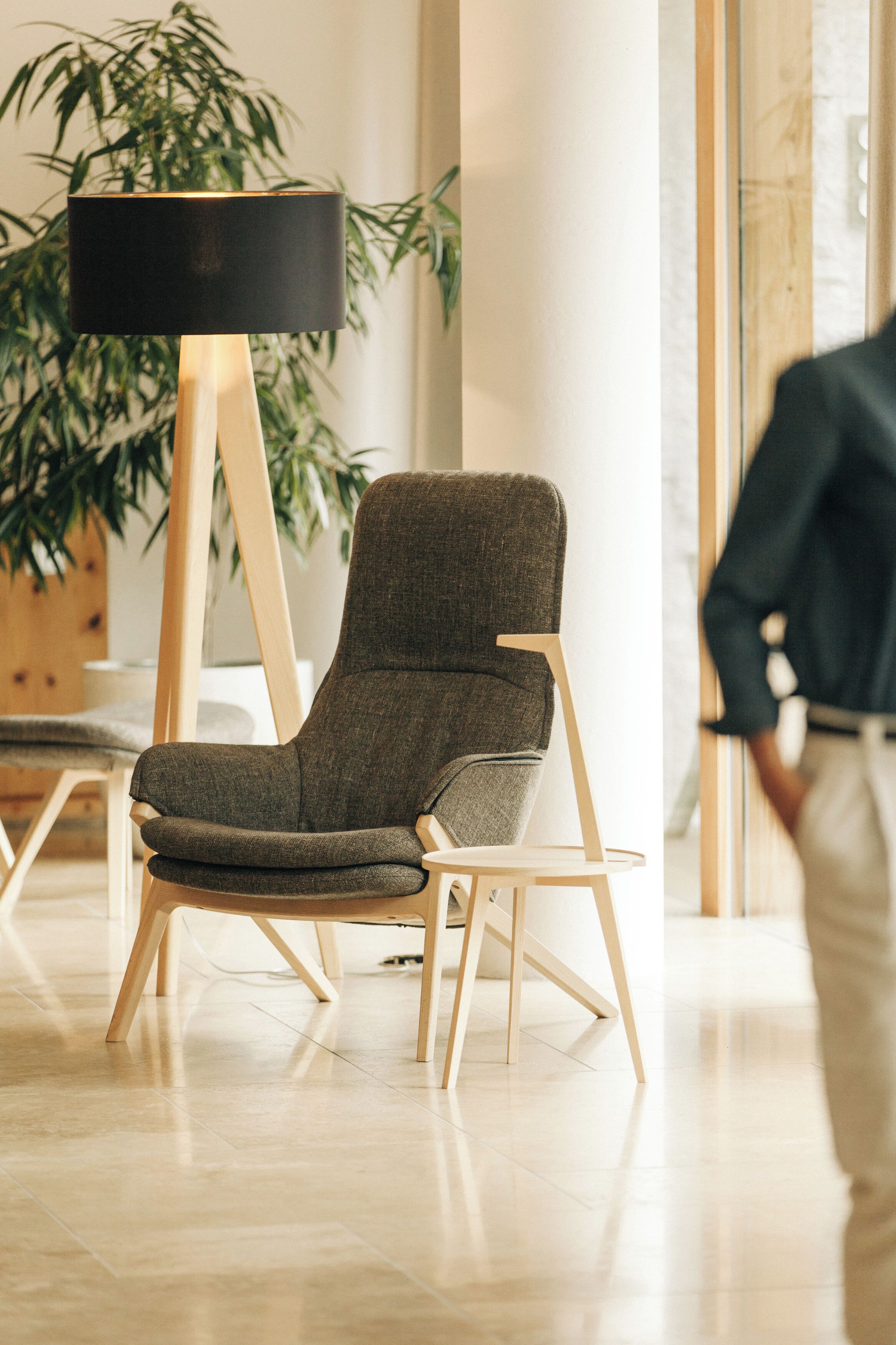 A modern chair with a dark fabric seat and backrest, wooden legs, and a black floor lamp with a wooden base in a well-lit room with a large plant in the background.