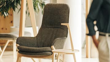 A modern chair with a dark fabric seat and backrest, wooden legs, and a black floor lamp with a wooden base in a well-lit room with a large plant in the background.