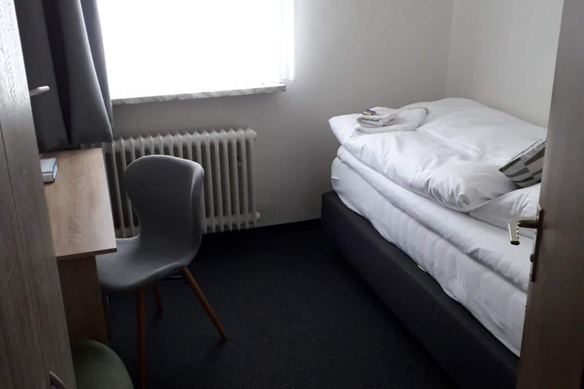 A small, single-bed room with a desk and chair, a radiator, and a window with curtains.