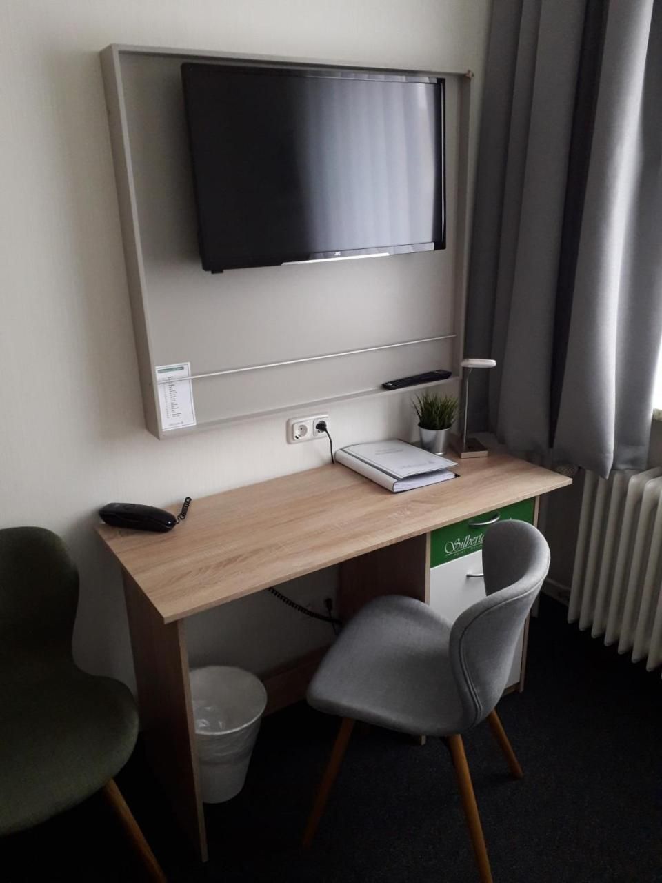 A hotel room with a wooden desk, a grey chair, a television mounted on the wall, a small potted plant, and a radiator.