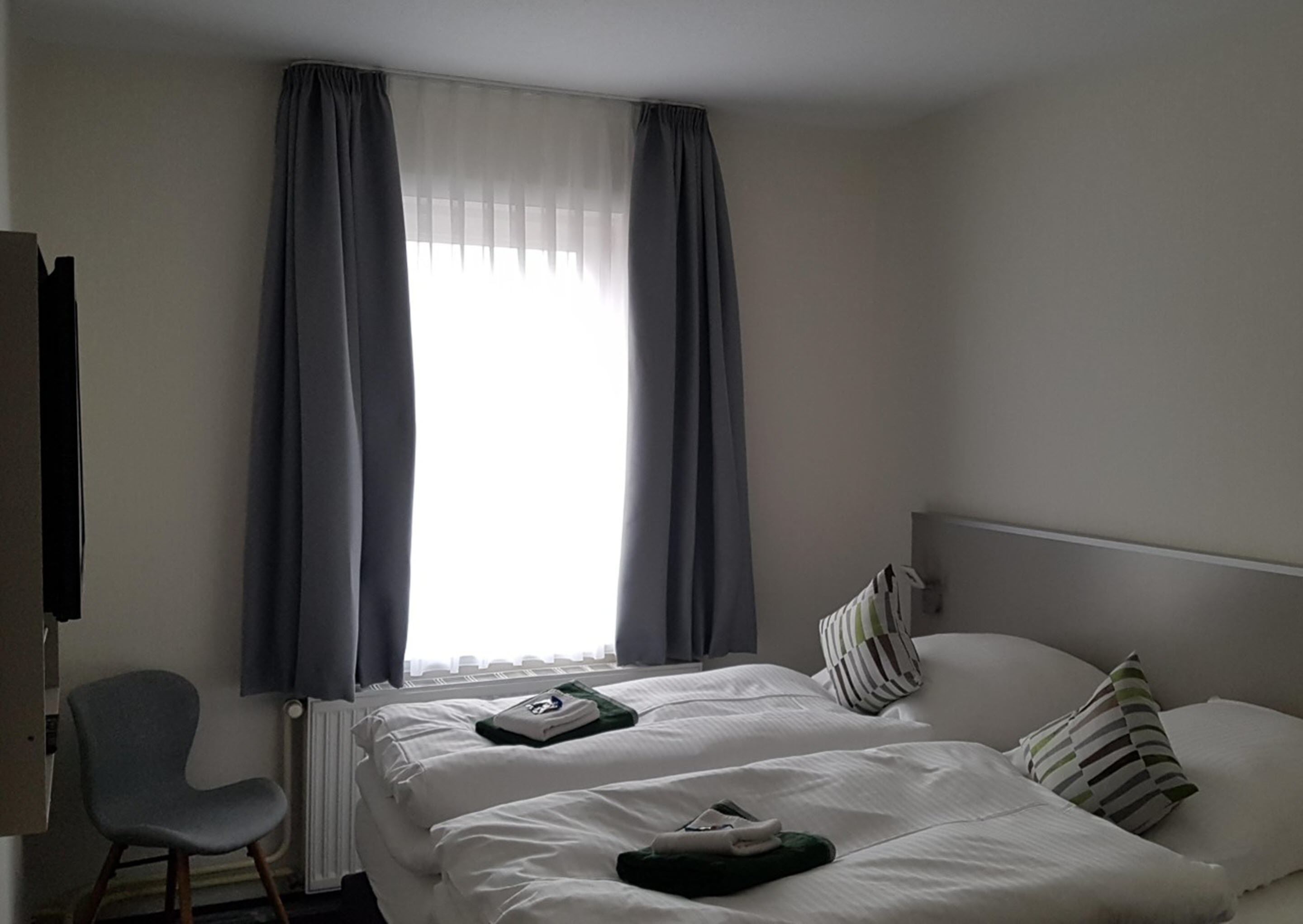 Quarto de hotel com cama, duas almofadas, uma cadeira, radiador e janela com cortinas.