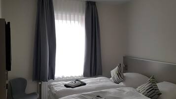 A hotel room with a bed, two pillows, a chair, a radiator, and a window with curtains.