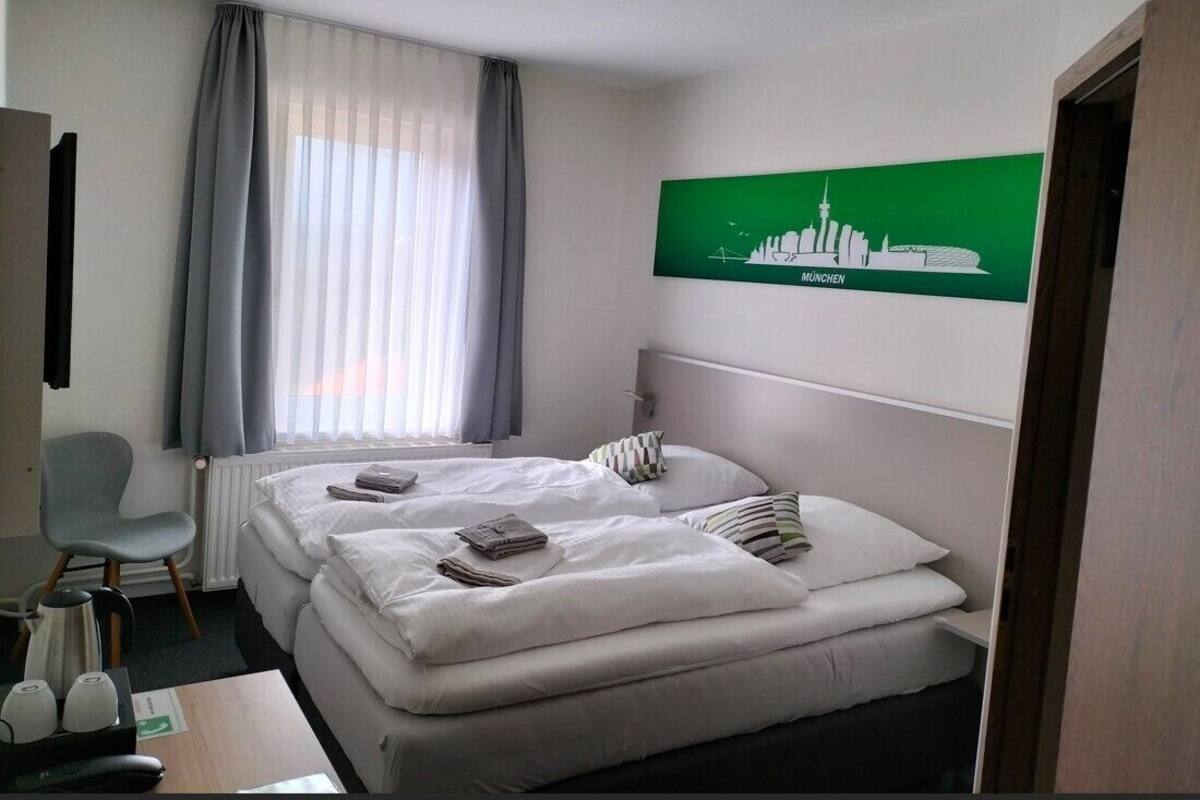 A hotel room with two beds, a chair, a radiator, a table with a kettle, and a cityscape picture on the wall.