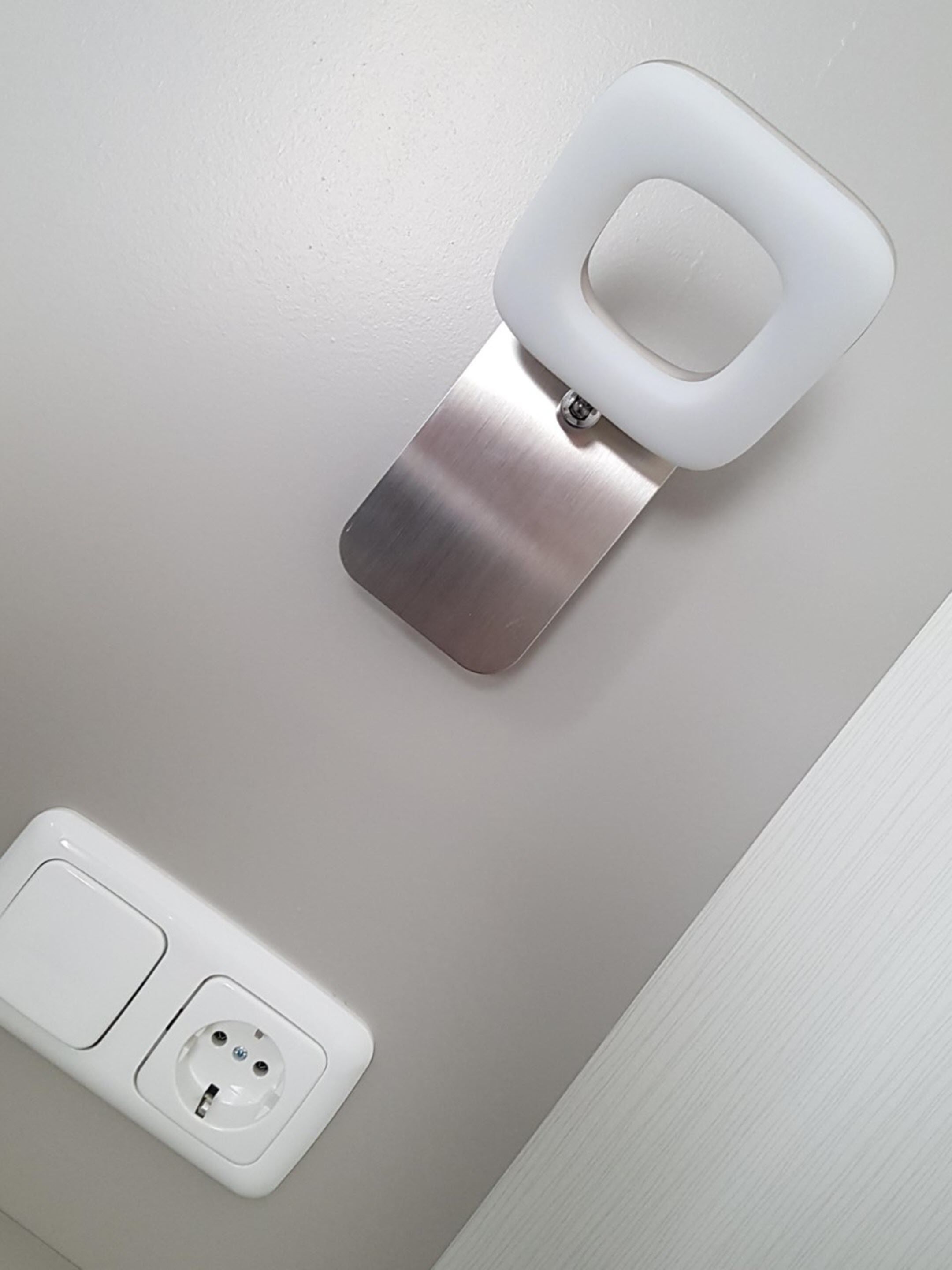 A wall-mounted toilet with a flush handle and a metal support bracket. Below it, there is a white electrical outlet with two sockets.