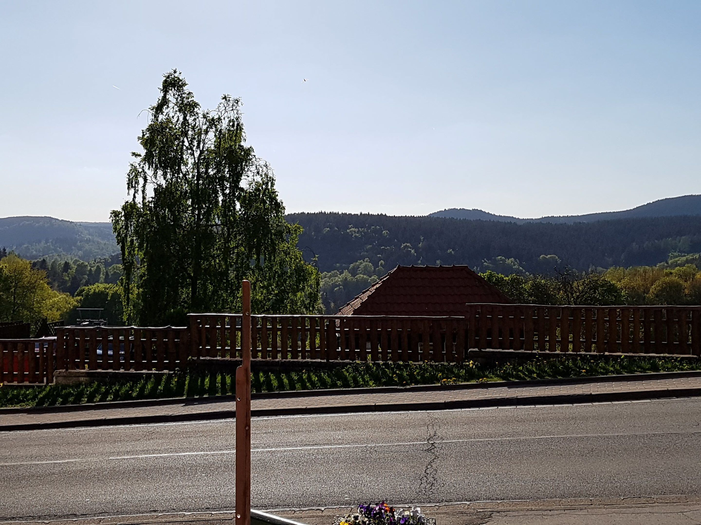 A view of a road with a wooden fence and a building with a red roof in the background.