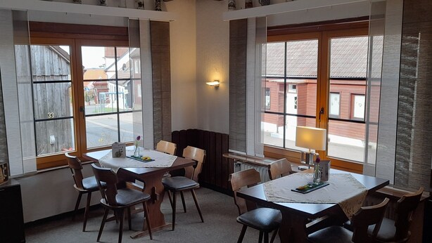 Un comedor con una mesa y sillas de madera, ventanas amplias con vista a la calle y un estante con objetos decorativos.