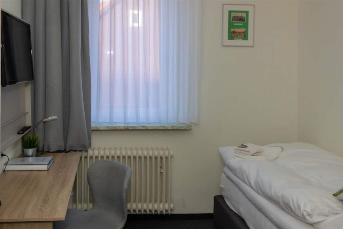 A small, tidy room with a bed, a desk, a chair, a radiator, and a window with curtains.
