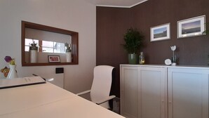Una oficina moderna con un escritorio blanco, una silla blanca, un espejo grande, un armario con dos cuadros enmarcados y plantas en macetas.
