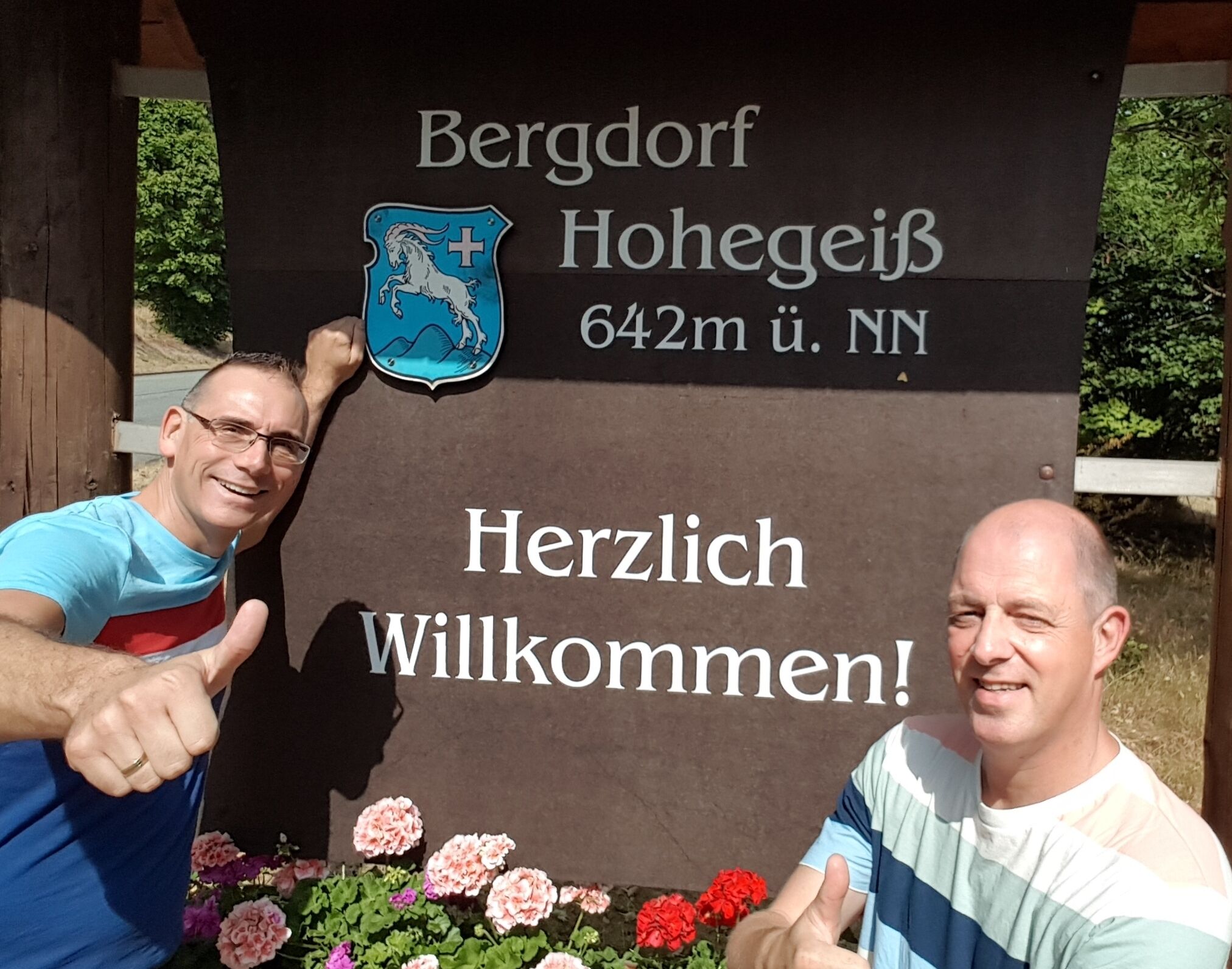Un letrero de bienvenida con un escudo y el texto 'Bergdorf Hohegeis 642m ü. NN' y '¡Bienvenido!'.