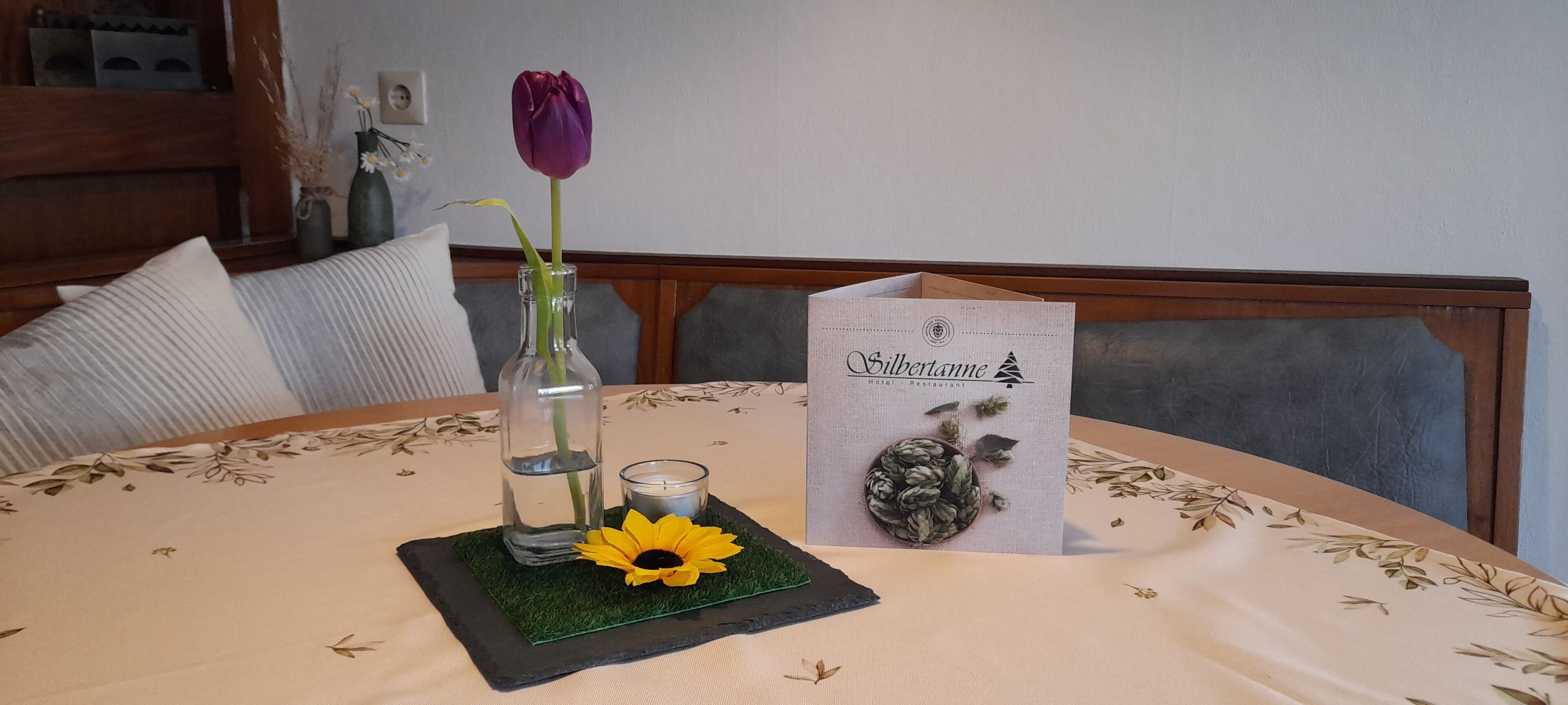 Una mesa con mantel floral, un florero con una flor morada, una vela y una tarjeta de menú.