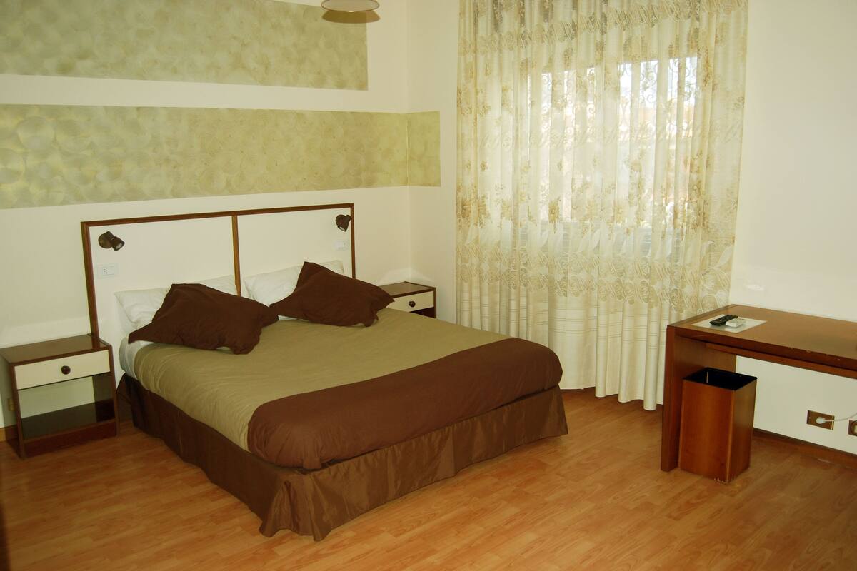 A bedroom with a bed, nightstands, a desk, and a trash bin.