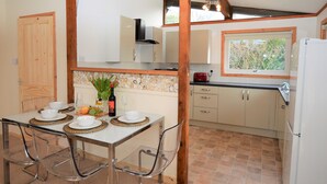 Cozinha com área de refeições, com mesa branca e cadeiras transparentes, painel de pedra e vigas de madeira.