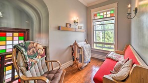 Ein gemütliches Zimmer mit einem Holzstuhl, einem roten Sofa, einer Holzhocker mit Dekorationen und einem Fenster mit Blick auf Grünflächen.
