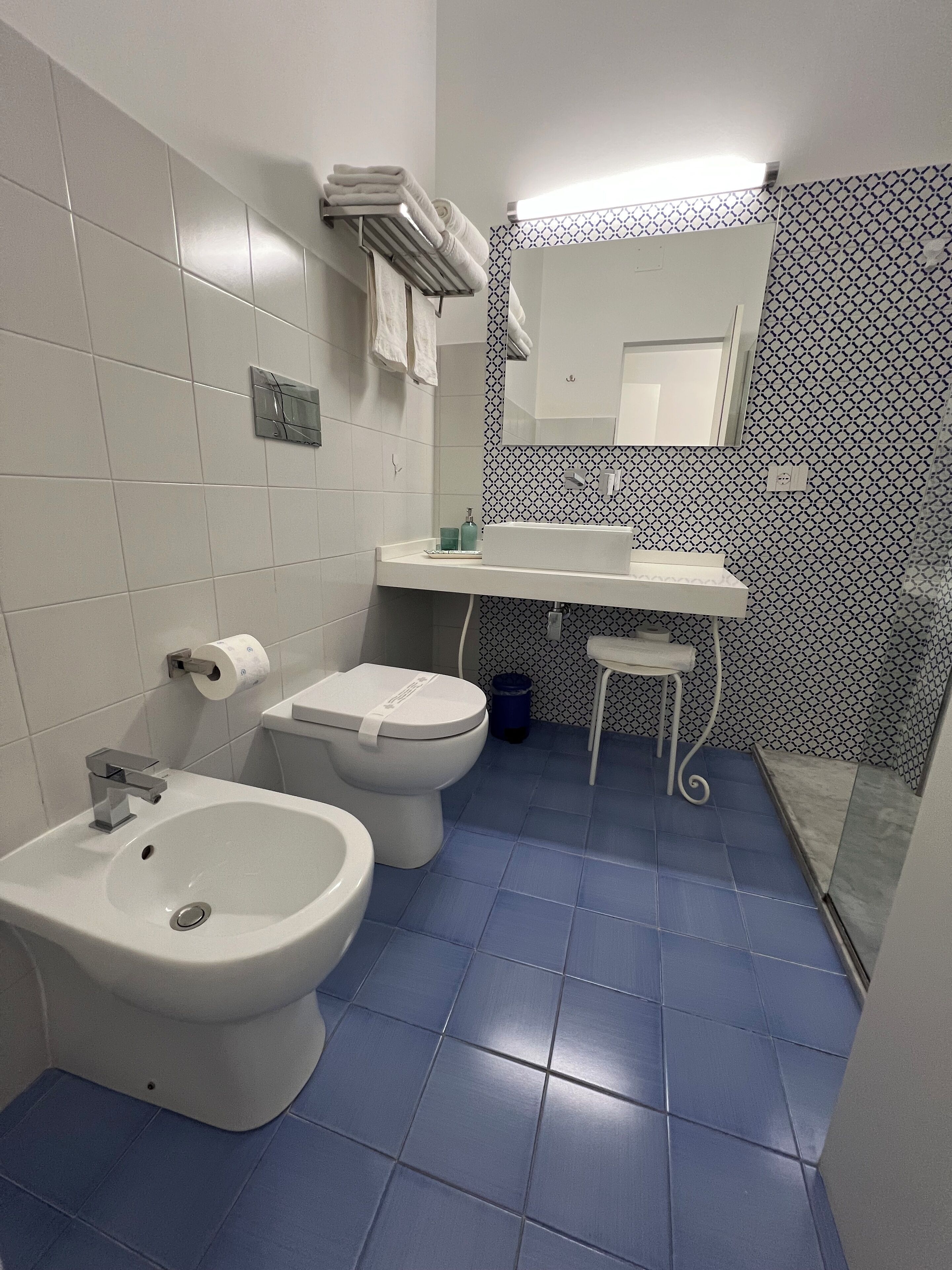 A bathroom with a white sink, a toilet, and a bidet. There is a mirror above the sink and a patterned wall behind it.