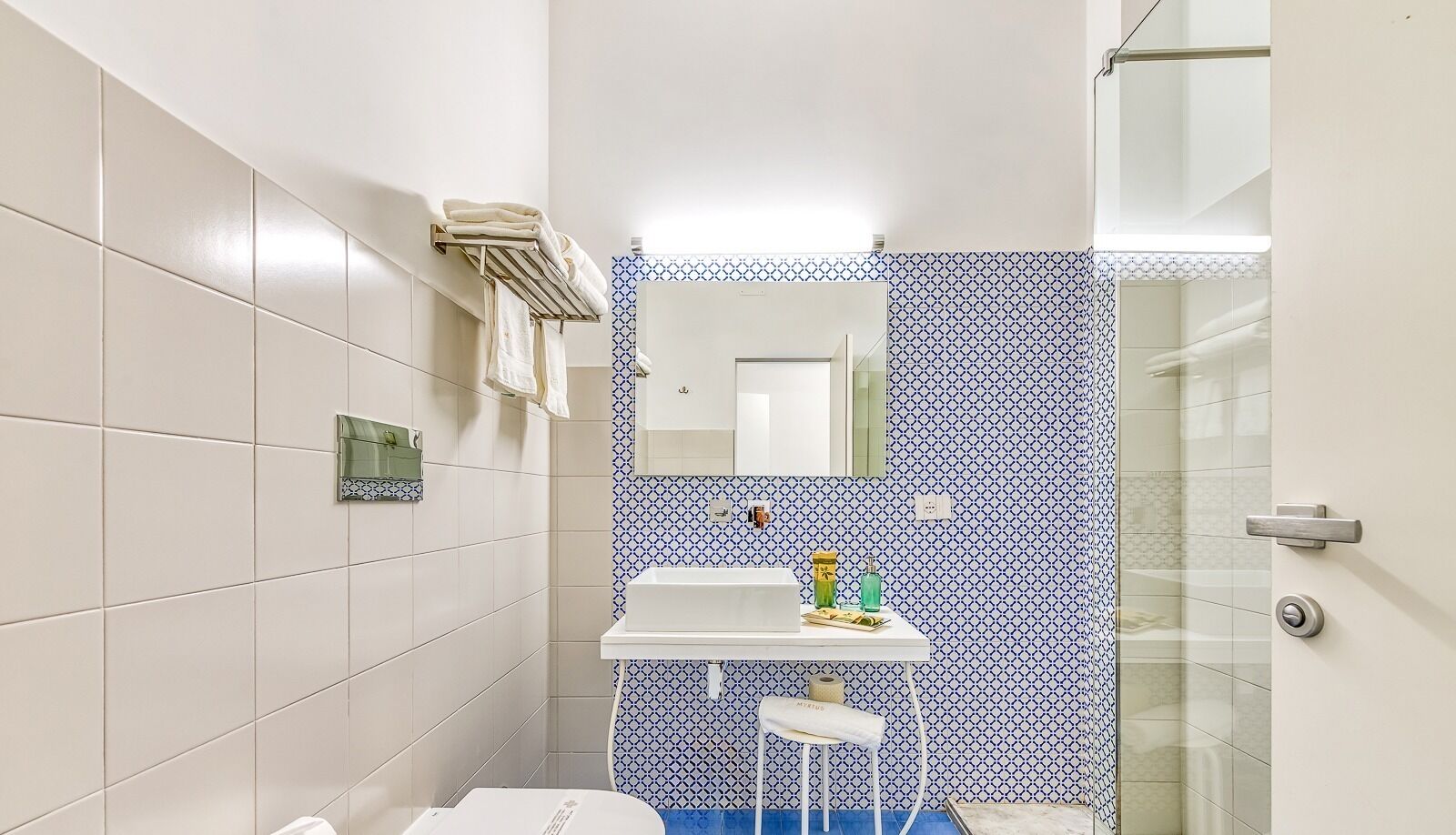 Un baño con baldosas blancas, un zócalo azul y blanco, un lavabo blanco, un espejo y un box de ducha de vidrio.
