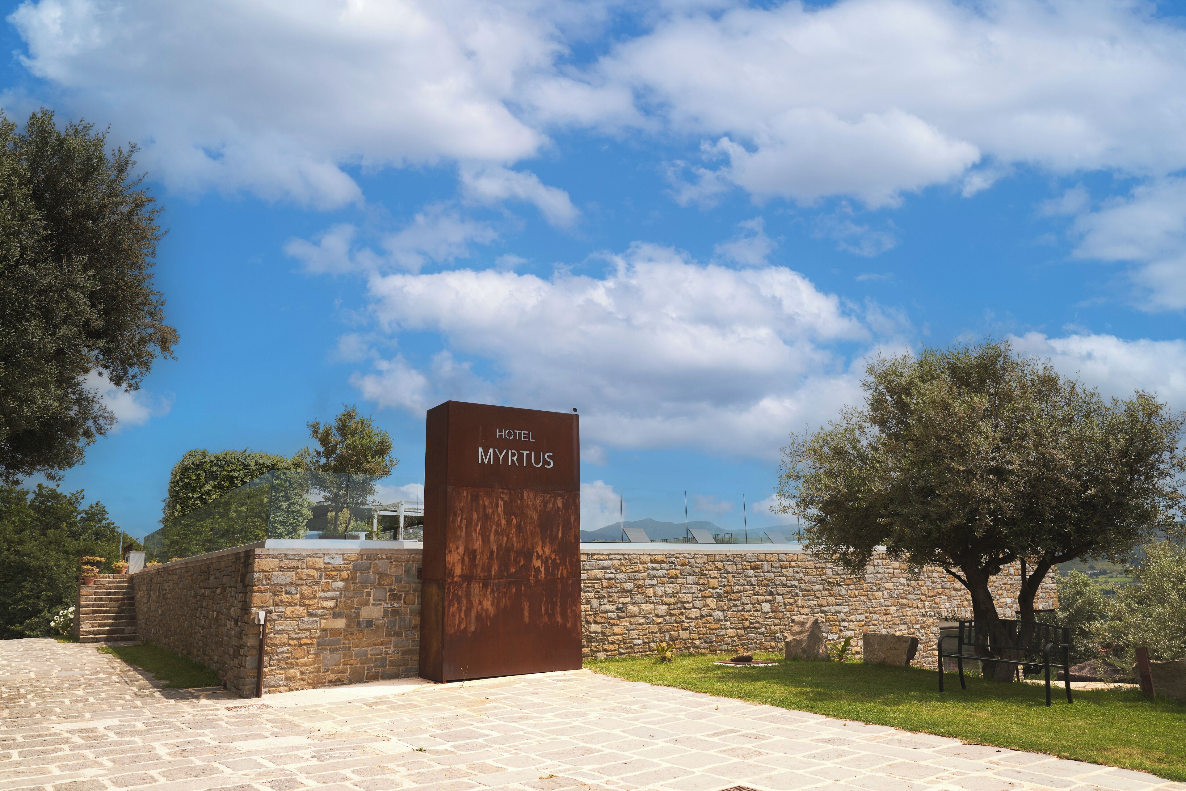 Un muro di pietra con un cartello che riporta "HOTEL MYRTUS" e un cielo azzurro con nuvole.