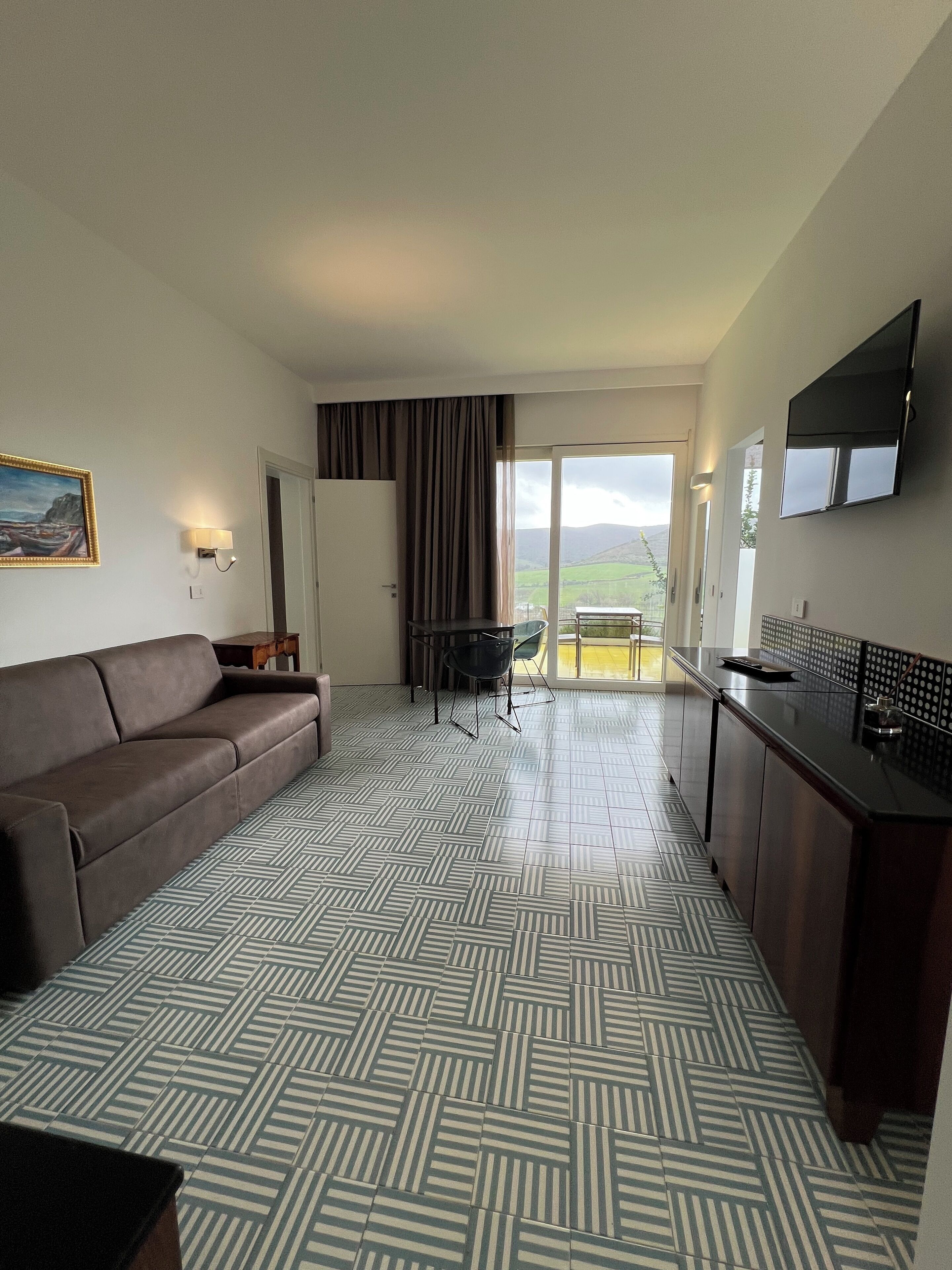 A modern hotel room with a sofa, dining table, and chairs, a kitchen area, and a balcony with a view.