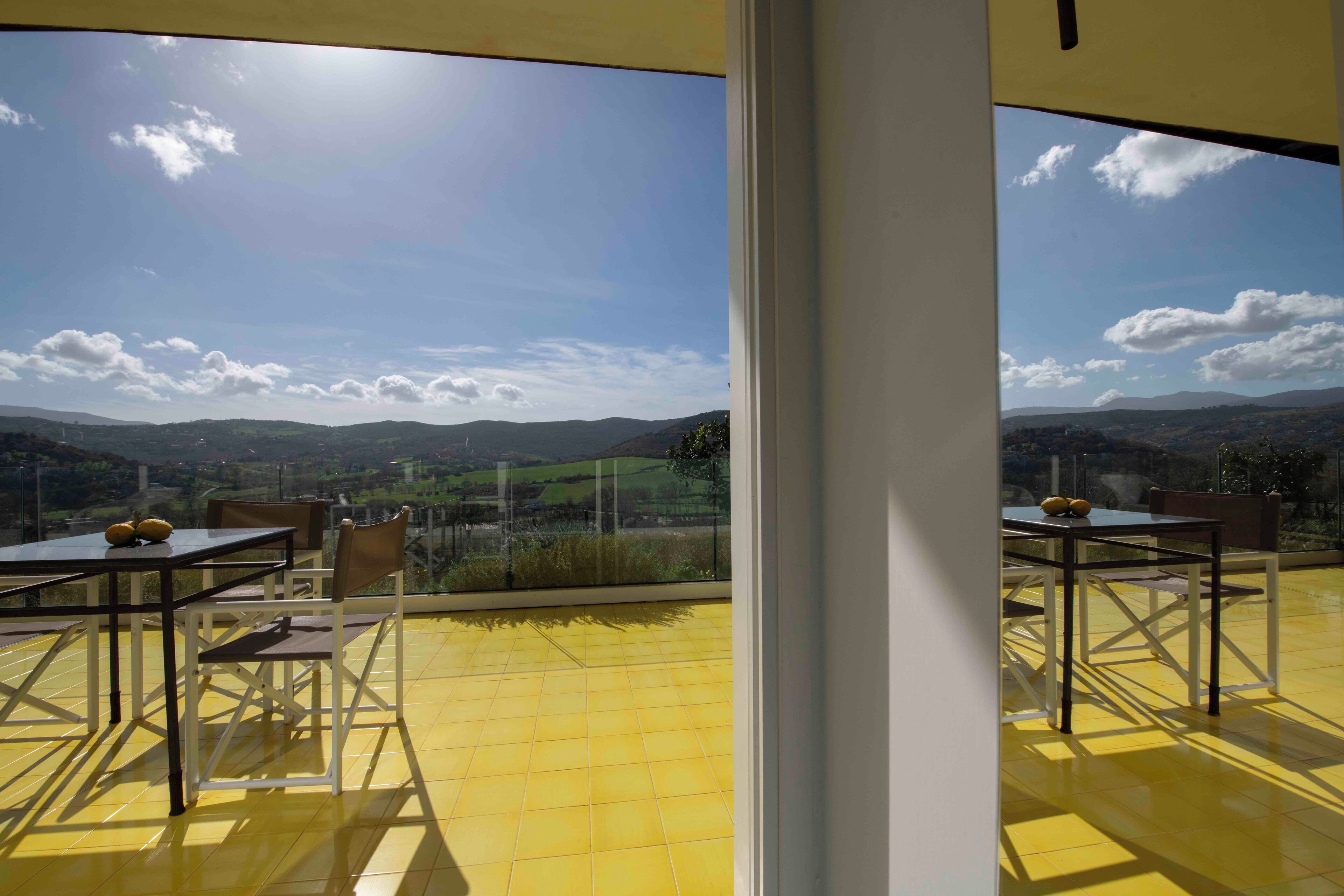 A terrace with yellow flooring, tables, and chairs, overlooking a scenic landscape with hills and a clear sky.