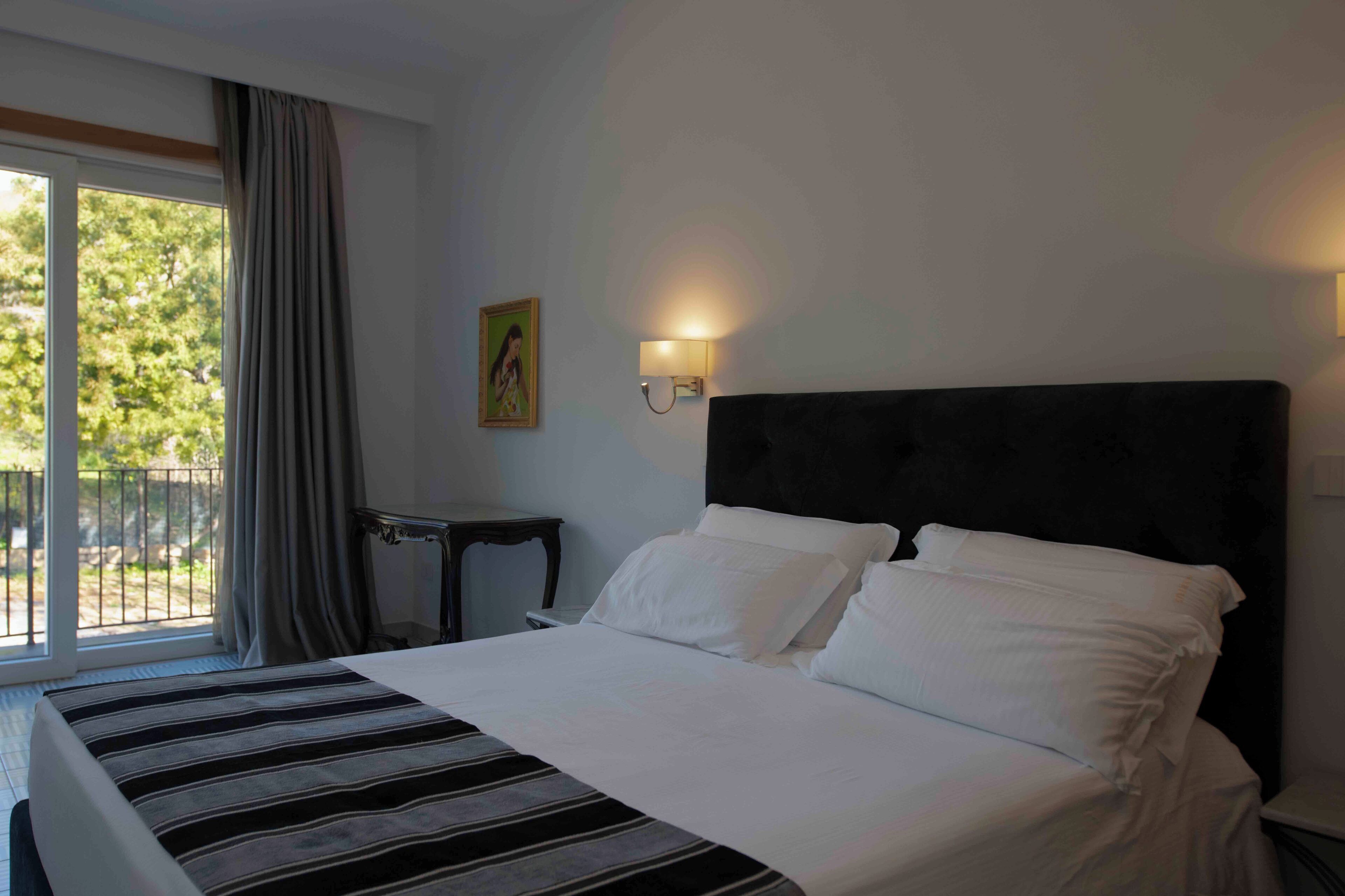 A hotel room with a large bed, a nightstand, a wall-mounted lamp, and a window with curtains.