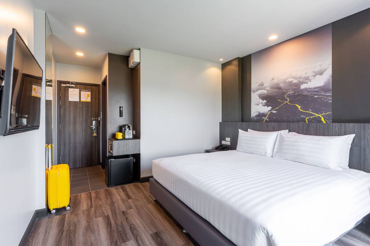 A hotel room with a large bed, a mirror, a small desk, a chair, a suitcase, and a wall-mounted TV.