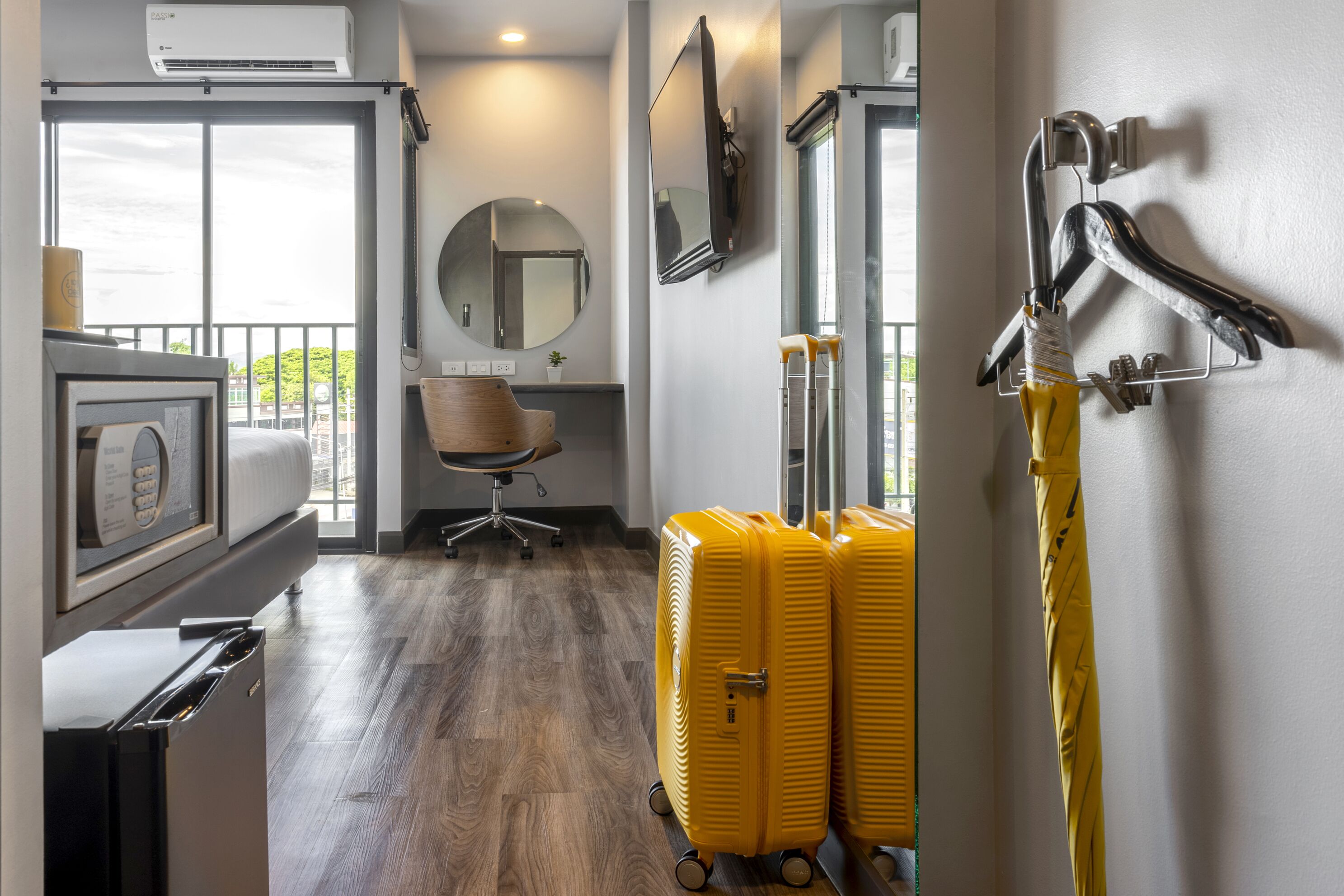 A hotel room with a large window, a desk, a chair, a mirror, and two yellow suitcases.
