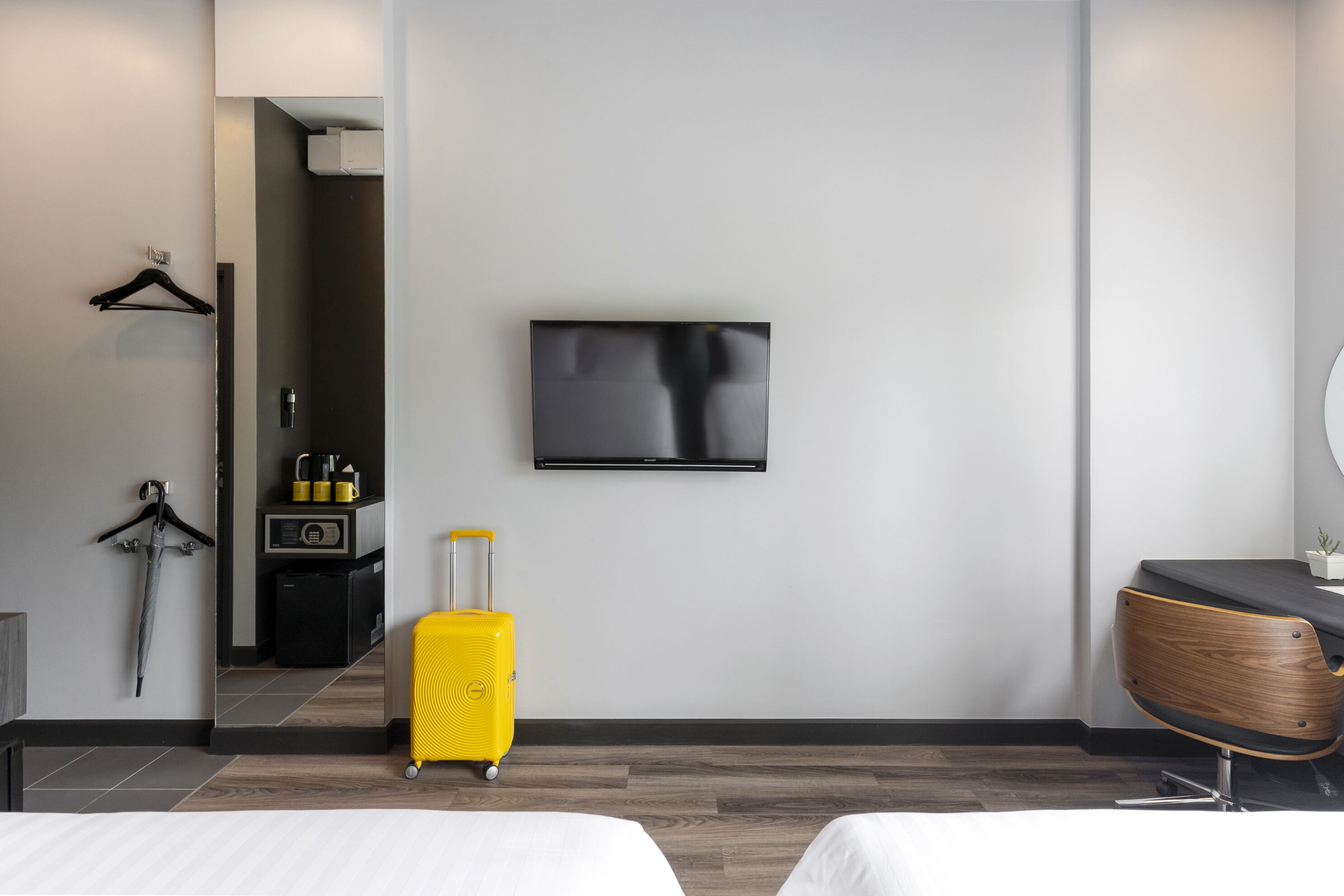 A modern hotel room with a bed, a TV mounted on the wall, a small desk with a chair, and a suitcase.