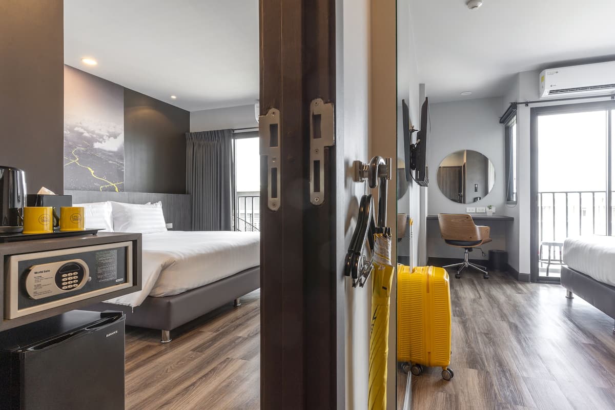 A hotel room with a bed, a desk with a computer, a chair, a suitcase, and a window with a view.