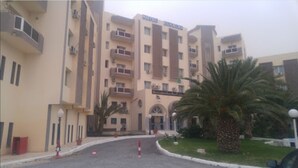 Een hotel met meerdere verdiepingen, een beige gevel, balkons op elke verdieping en een centrale ingang met een palmboom op de voorgrond.