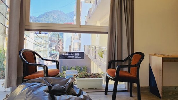 A room with a bed, two chairs, a desk, and a view of a cityscape through large windows. - Chakrah by Hermitage (Narendranagar)
