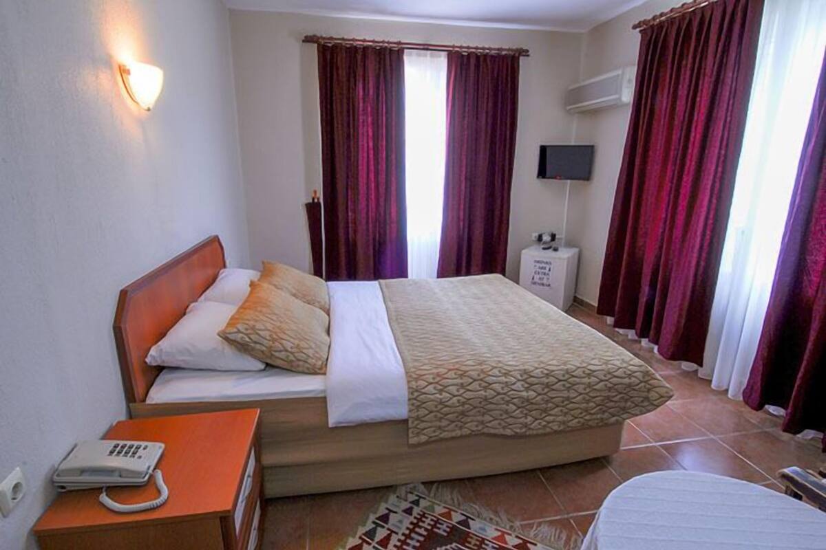 A bedroom with a bed, a bedside table with a phone, a TV on the wall, and curtains.
