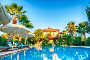 A swimming pool with lounge chairs and umbrellas, surrounded by palm trees and a large building in the background. - Göcek Alize Hotel (Fethiye)