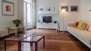 Una sala de estar moderna con un televisor de pantalla plana, una mesa de centro de vidrio, un sofá con cojines coloridos y un mueble de entretenimiento blanco con objetos decorativos.