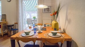 Un comedor con una mesa de madera dispuesta para cuatro personas, sillas, un reloj en la pared y una ventana con toldo a rayas.