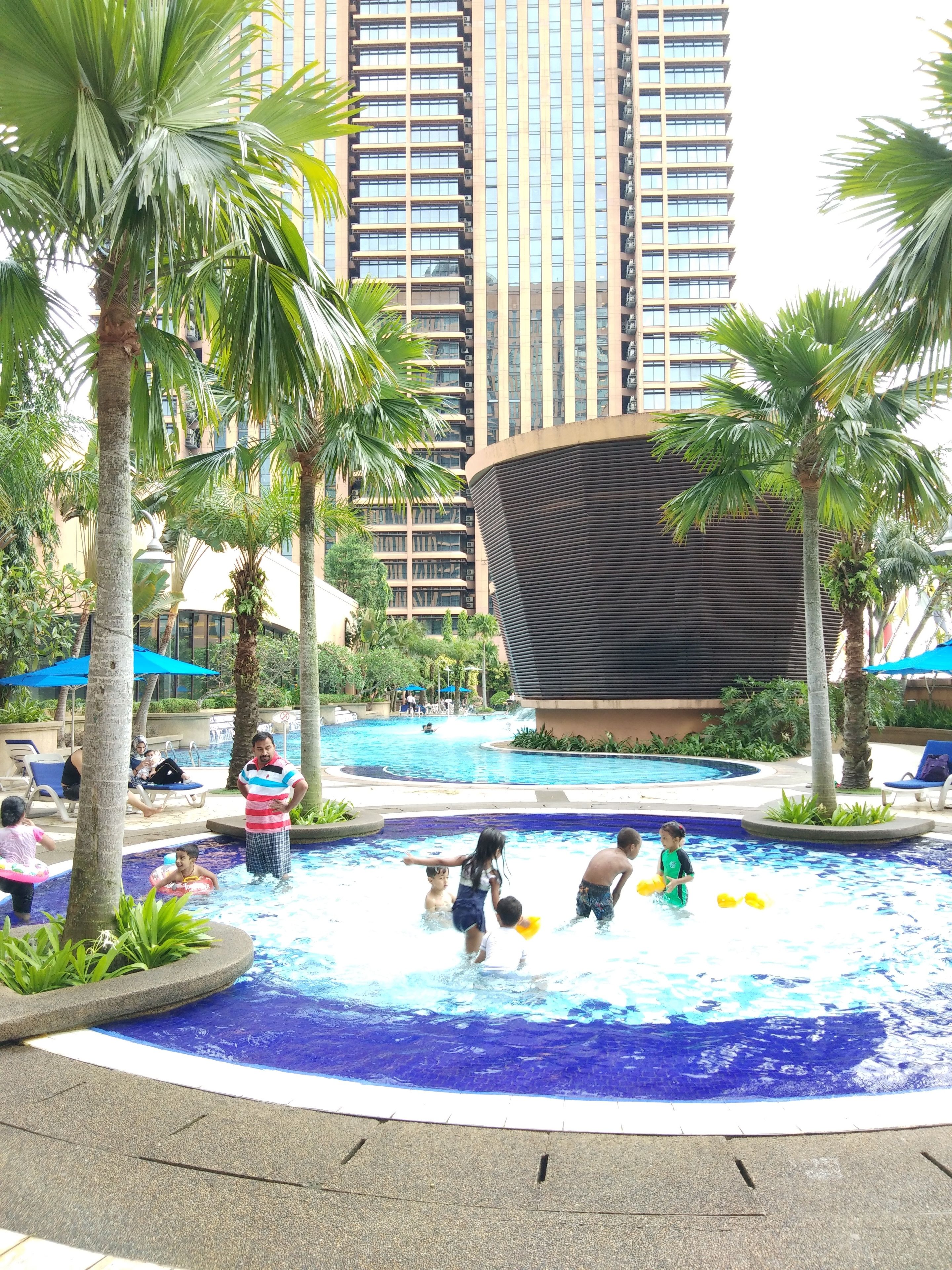 A swimming pool area with palm trees, a children's play area, and a modern building in the background.