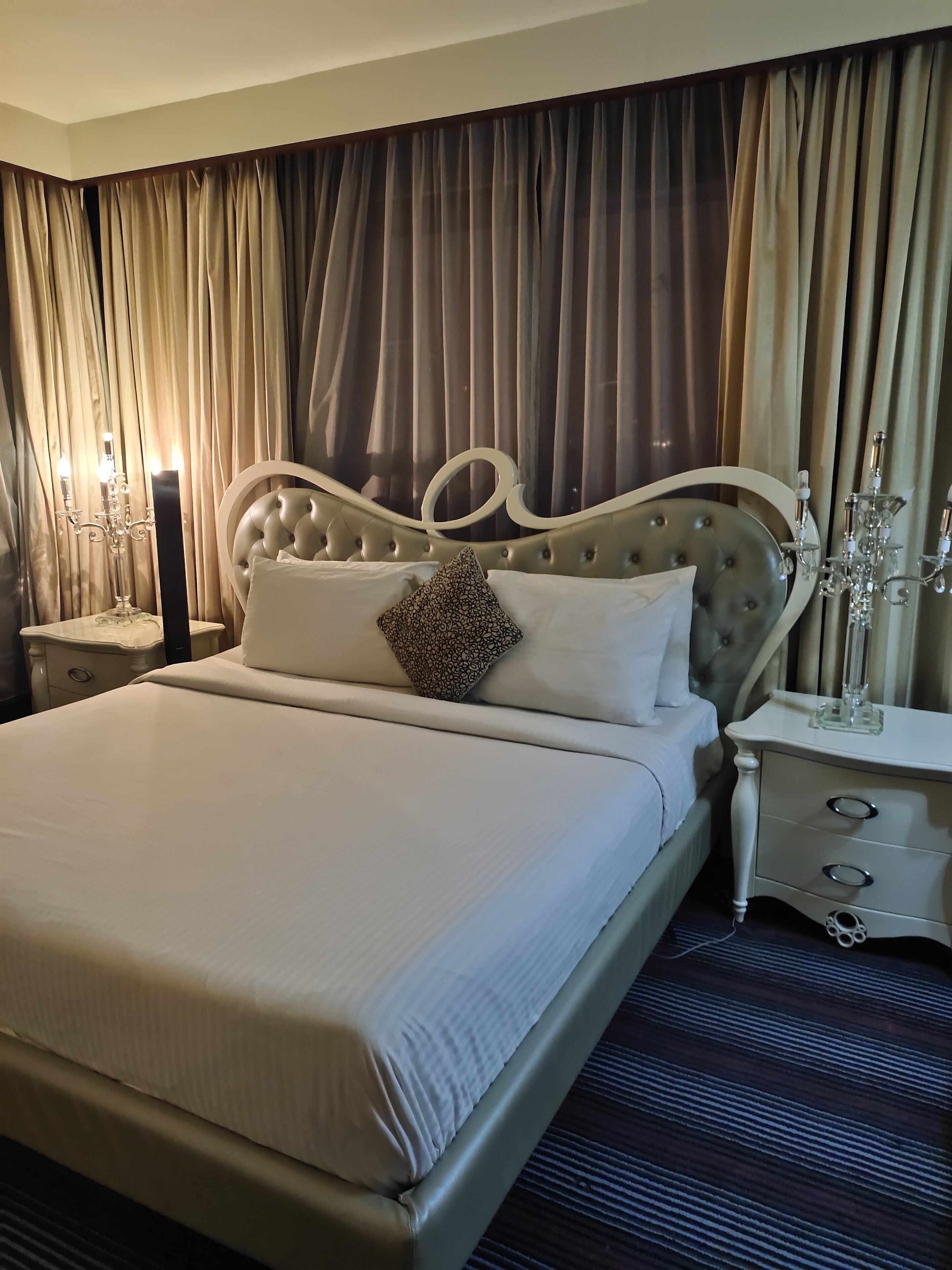 A neatly made bed with a tufted headboard, a bedside table with a lamp, and a large window with curtains.
