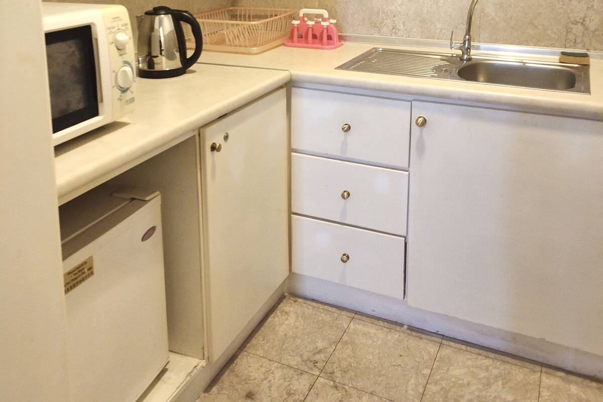 A compact kitchen with white cabinets, a microwave, a kettle, a sink, and a small refrigerator.