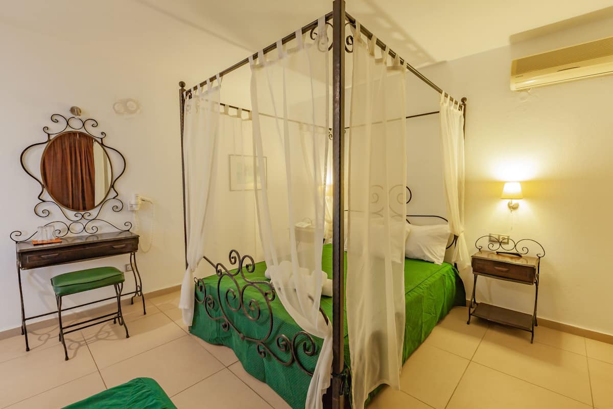 A bedroom with a four-poster bed, a bedside table, a desk with a chair, and a mirror.