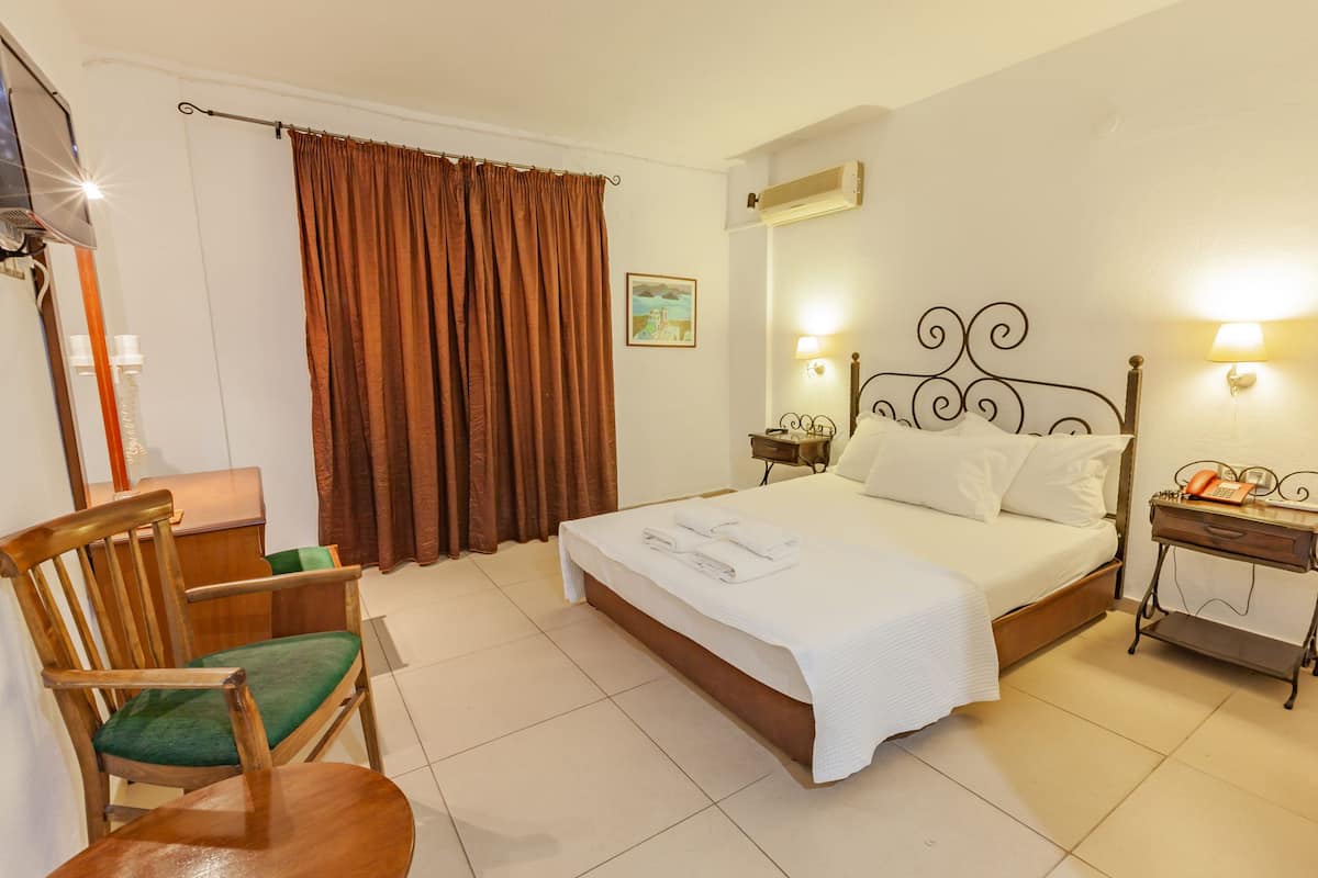 A hotel room with a bed, a chair, a bedside table, a lamp, and a wall-mounted air conditioner.