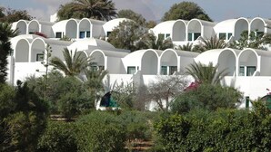 Uma fileira de edifícios brancos com janelas e varandas em arco, cercada por vegetação e palmeiras.