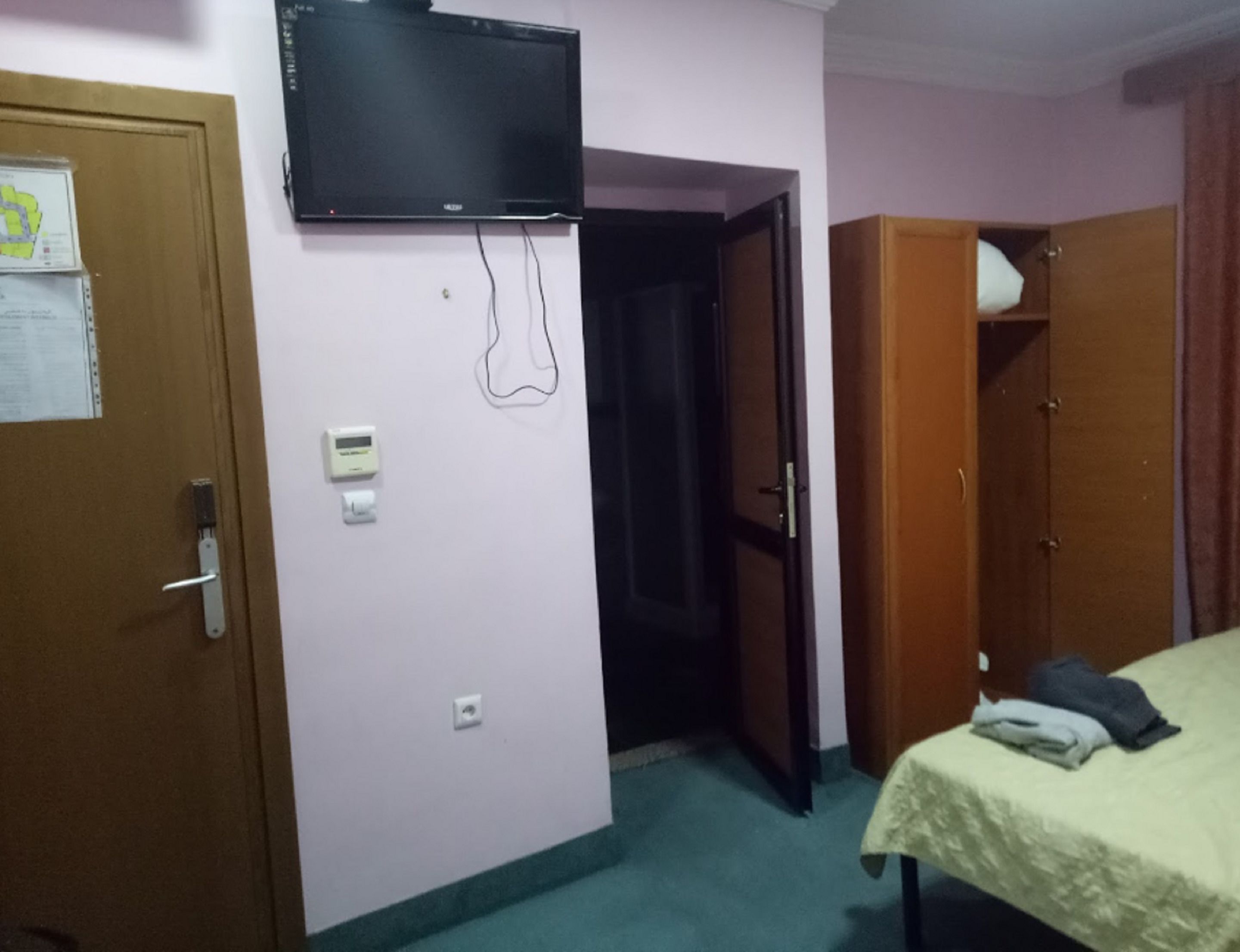 Habitació amb un llit, una televisió penjada a la paret, una porta amb un tirador i una presa elèctrica petita.